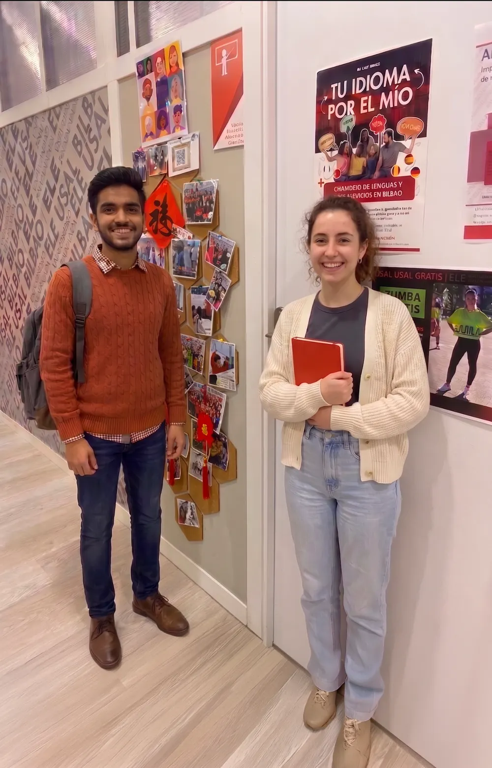 Dos estudiantes internacionales sonríen en el pasillo de la escuela de español ELE USAL Bilbao. Dos llevan mochilas y una estudiante sostiene una libreta roja. En las paredes se ven carteles sobre actividades culturales y aprendizaje de idiomas, reflejando un ambiente multicultural y acogedor.