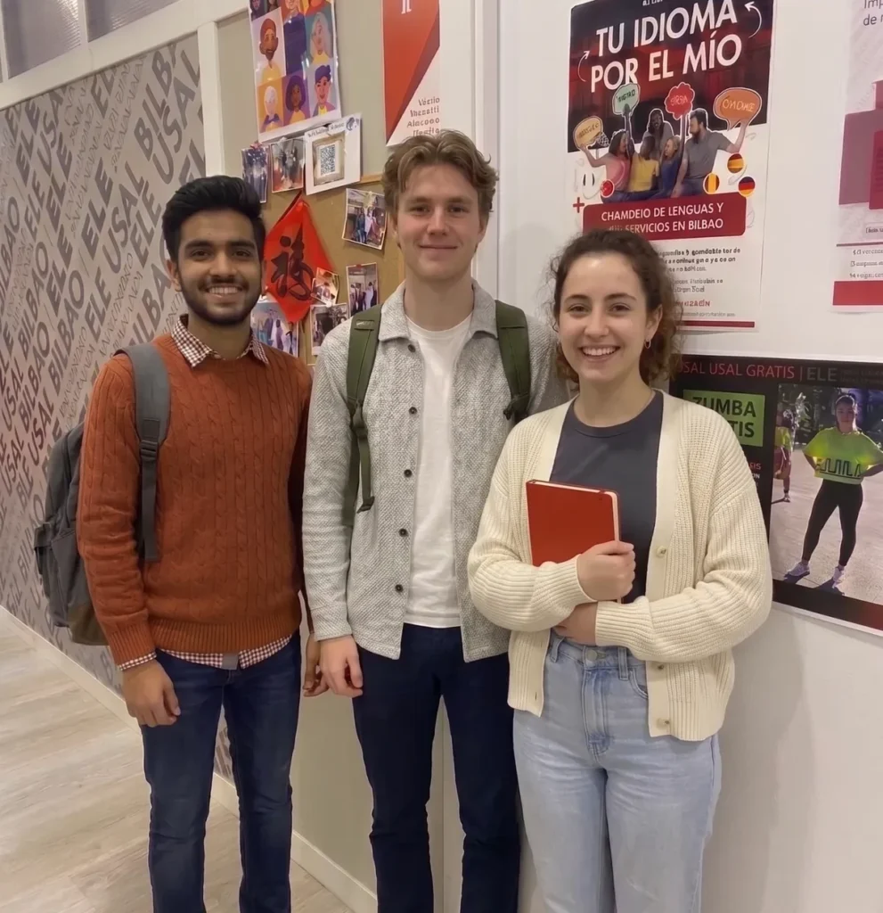 Tres estudiantes internacionales sonríen en el pasillo de la escuela de español ELE USAL Bilbao. Dos llevan mochilas y una estudiante sostiene una libreta roja. En las paredes se ven carteles sobre actividades culturales y aprendizaje de idiomas, reflejando un ambiente multicultural y acogedor.